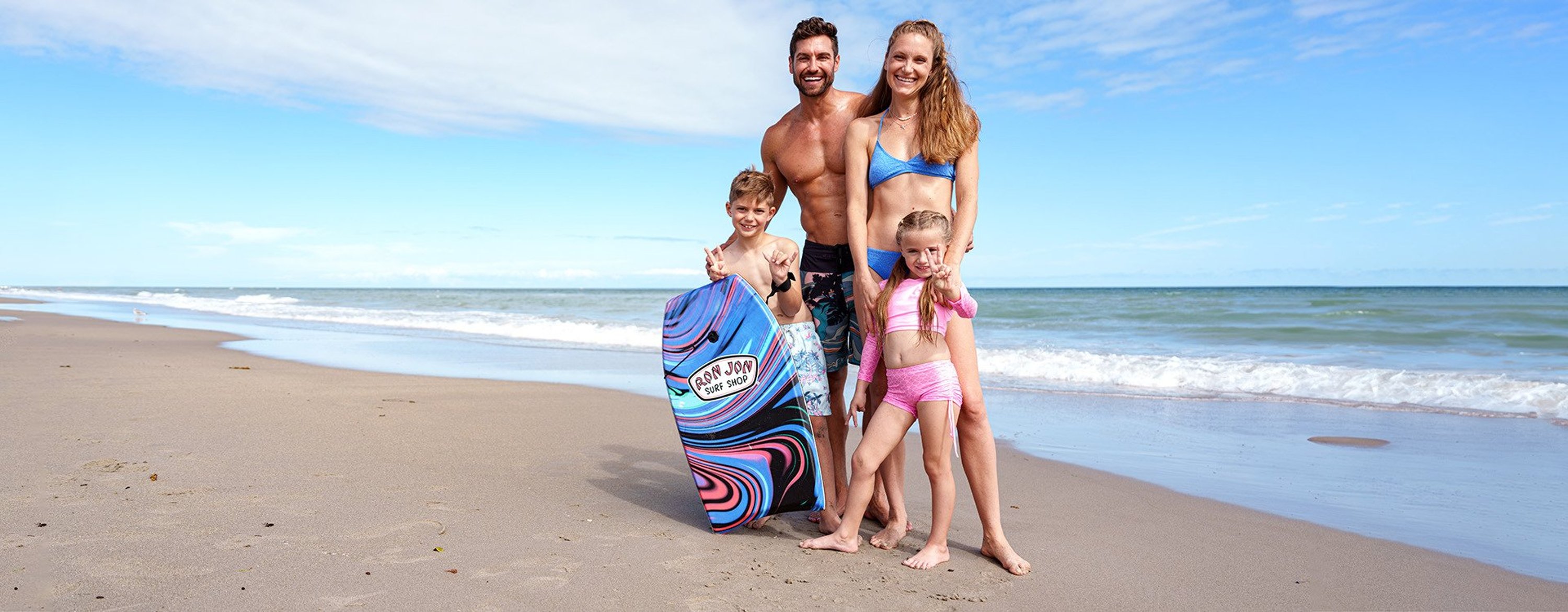 family standing on the beach at the waters edge with the youngest holding a bodyboard